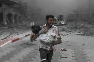 A paramedic holding a child in his running in the middle of a destructed area.